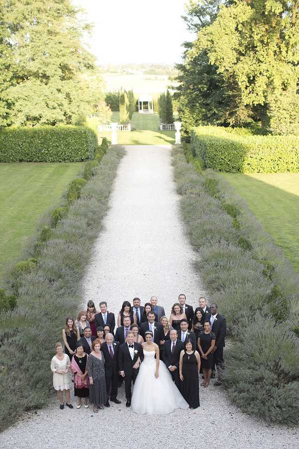 A group portrait of approximately 25-30 wedding guests and the couple posed on a formal gravel garden allee lined with lavender borders and neatly trimmed low hedges, with cypress trees and a white classical pergola or pavilion visible in the distance. The bride wears a strapless white ballgown with a full skirt, and the groom is in a black tuxedo with a white boutonniere; guests are dressed in formal attire including black dresses, dark suits, and a few lighter-colored outfits. The shot is taken from an elevated angle looking down the length of the allee, giving a wide, slightly aerial perspective that emphasizes the formal symmetry of the garden design. The setting appears to be the grounds of a French chateau with classic formal French garden landscaping. Potential venue feature image.
