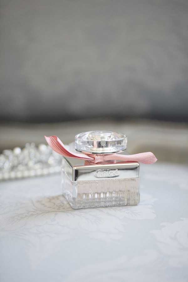 Close-up detail shot of a Chloé perfume bottle placed on a white embroidered surface during a bridal getting-ready scene. The clear glass bottle with a silver mirrored band features the brand name embossed in silver lettering and is tied with a dusty pink grosgrain ribbon bow around the neck. A pearl bracelet or necklace is partially visible and softly blurred in the left background, suggesting a styled bridal accessories flat lay. The image uses a shallow depth of field with a soft, neutral grey background.