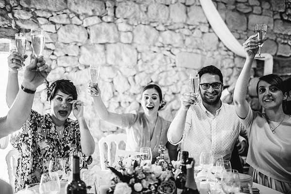 Black-and-white image of a champagne toast during a wedding reception, with four guests in the foreground raising their flutes and glasses with animated, joyful expressions. The setting is an indoor venue with exposed stone walls, likely a French chateau or barn conversion, with what appears to be a fabric drape visible in the upper right corner. The table in the foreground holds wine bottles, glassware, and a floral centerpiece with what appear to be roses and mixed blooms. The composition is a mid-range shot capturing the energy and spontaneity of the moment.