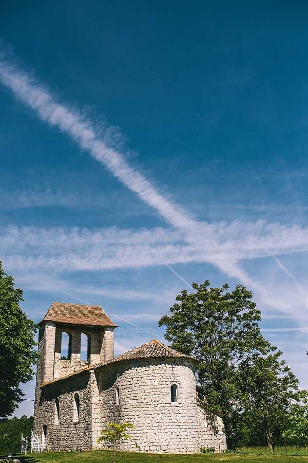 Wide-angle exterior shot of a small Romanesque stone chapel with a square bell tower and terracotta-tiled roof, photographed from ground level looking up. The chapel features a rounded apse constructed from pale limestone blocks with narrow arched windows. A row of white chairs is partially visible to the left of the building, indicating an outdoor ceremony setup. The composition is a low-angle wide shot with the architecture positioned in the lower third of the frame. Potential venue feature image.