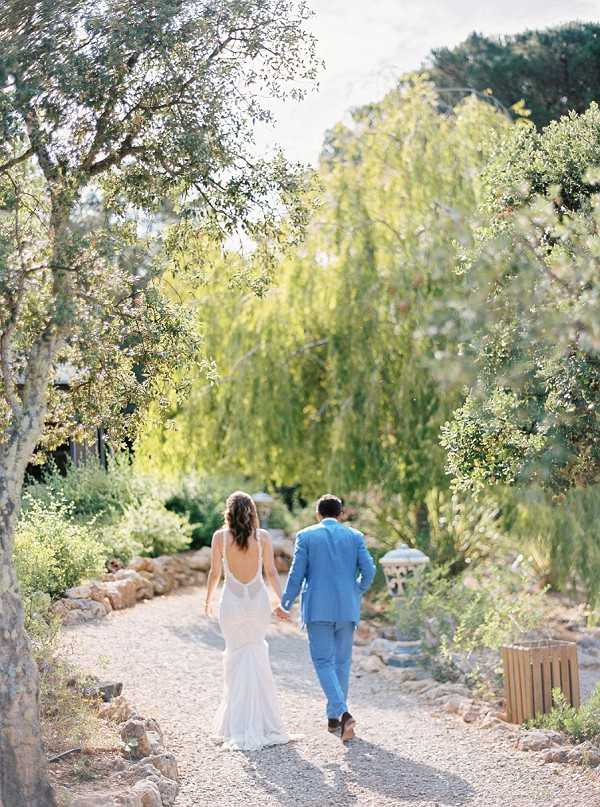 A couple walks hand-in-hand away from the camera along a gravel path through a lush garden setting, captured in a wide portrait shot. The bride wears a white sleeveless gown with a low open back and a fitted silhouette, her dark hair worn down in loose waves. The groom is dressed in a light blue suit with no tie. The garden path is bordered by rockery edging and dense Mediterranean-style plantings, with a weeping tree creating a natural canopy in the background, giving the scene a relaxed, boho-garden atmosphere.