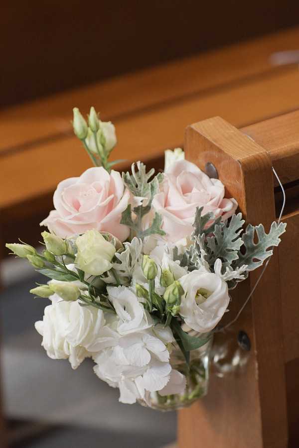 Close-up detail shot of a pew end floral arrangement hanging from a wooden church pew in a clear glass jar. The small bouquet features blush pink roses, white lisianthus blooms and buds, white carnations, and dusty miller foliage with its characteristic silver-grey lobed leaves. The soft blush and white color palette against the warm honey-toned wood of the pew creates a classic, understated ceremony decor look. The arrangement is tied to the pew end with a thin clear wire or monofilament.