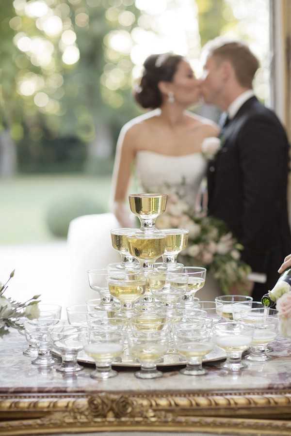 A champagne tower of coupe glasses filled with pale golden champagne is the sharp focal point in the foreground, arranged in a pyramid on a silver tray atop a marble-topped table with a gilt frame. In the softly blurred background, the bride and groom share a kiss; the bride wears a strapless white gown with her dark hair pinned up, and the groom wears a dark suit with a light blue tie. A lush arrangement of blush and white flowers with greenery is visible beside them, and the setting appears to be an interior space with large windows or doors opening onto a green outdoor garden. The composition is a close-up detail shot with intentional depth-of-field separating the champagne tower from the couple behind it.