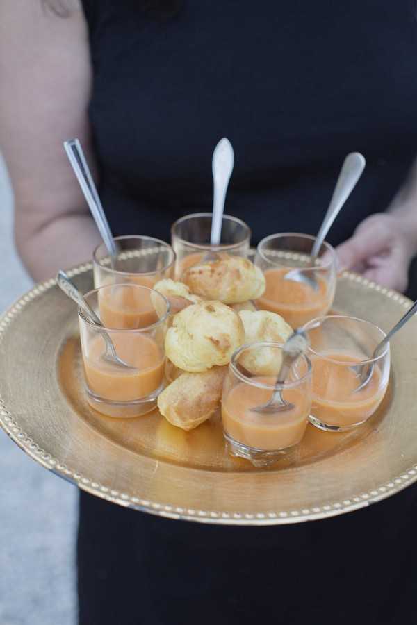 A close-up shot of a catering staff member dressed in black holding a round gold tray during what appears to be a cocktail hour. The tray is arranged with seven small clear glasses filled with an orange-toned soup or velouté, each with a small silver spoon, surrounding a cluster of golden choux pastry puffs in the center. The presentation is neat and the gold tray adds a warm metallic accent to the passed appetizer service.