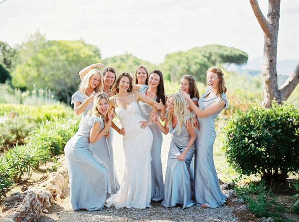 A bridal party portrait taken outdoors in a garden or estate setting, likely in the south of France, with Mediterranean vegetation visible in the background. The bride stands at the center wearing a fitted white lace spaghetti-strap gown, surrounded by eight bridesmaids in matching pale silver-blue satin cowl-neck floor-length dresses. The group is laughing and posing playfully, with several bridesmaids making fun gestures toward the camera. The image is a wide group portrait shot in bright natural daylight with a soft, warm tone consistent with film photography.