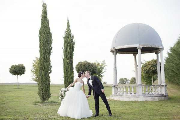 A couple shares a kiss during outdoor wedding portraits on the grounds of what appears to be a French estate or chateau. The bride wears a full-skirt white ballgown with a strapless silhouette and carries a bouquet of white and greenery flowers, while the groom is dressed in a classic black tuxedo with bow tie. They are positioned in front of a stone classical rotunda structure with a domed roof, columns, and balustrade railing, flanked by tall cypress trees and a trimmed lollipop-shaped topiary. The composition is a wide portrait shot with soft, overcast natural lighting giving a muted, airy tone to the image. Potential venue feature image.