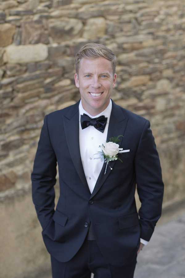 A portrait of the groom standing outdoors in front of a rustic stone wall. He is wearing a navy tuxedo with a white dress shirt, a black velvet bow tie, and a boutonniere featuring a blush rose with small greenery accents. He stands with both hands in his pockets and is smiling directly at the camera. The shot is a mid-length portrait with a classic, formal styling aesthetic.