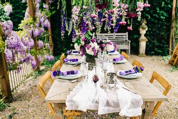 An outdoor reception tablescape styled in a garden setting, photographed as a wide-angle detail shot. A rustic wooden farm table with natural wood folding chairs is set for approximately eight guests on a gravel surface, with a white lace table runner running down the center. Place settings include white plates, silver glassware, and deep purple folded napkins. Centerpieces feature arrangements of ivory and purple blooms, including what appear to be peonies and lilacs, in dark vases alongside silver candle holders. A dramatic floral installation hangs overhead, composed of cascading purple delphiniums, deep burgundy blooms, and greenery. The overall decor palette is purple, burgundy, ivory, and silver against natural wood, creating a garden-romantic aesthetic. A white wooden bench and ornamental stone pedestal are visible in the background.