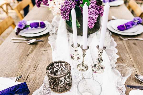 A close-up detail shot of a wedding reception tablescape set on a rustic wooden farm table. The centerpiece features a low arrangement of purple and white lilacs in a dark glass vessel, flanked by two white taper candles in silver candlestick holders. The table is dressed with a white lace runner, and place settings include white plates with purple napkins tied with purple ribbon or bands. Additional decorative elements include a dark metal filigree votive candle holder and a clear glass tumbler. The overall decor palette is purple, white, and silver with a vintage-romantic styling theme.