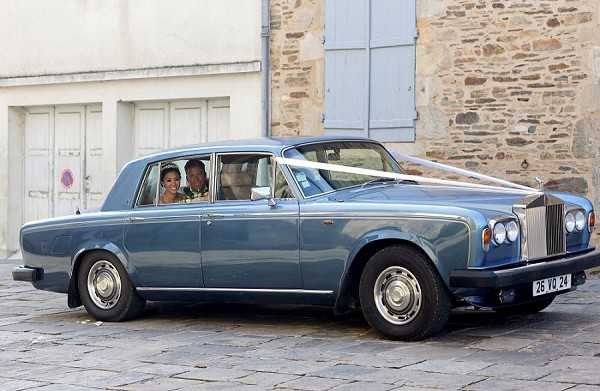 A bride and groom are seated inside a vintage steel-blue Rolls-Royce Silver Shadow, decorated with white ribbon across the hood, suggesting a wedding car transfer between venues. The vehicle has a French license plate and is parked on a cobblestone surface in front of a stone building with pale blue shuttered doors. The couple is visible through the rear and side windows. Wide shot composition capturing the full length of the classic luxury car.