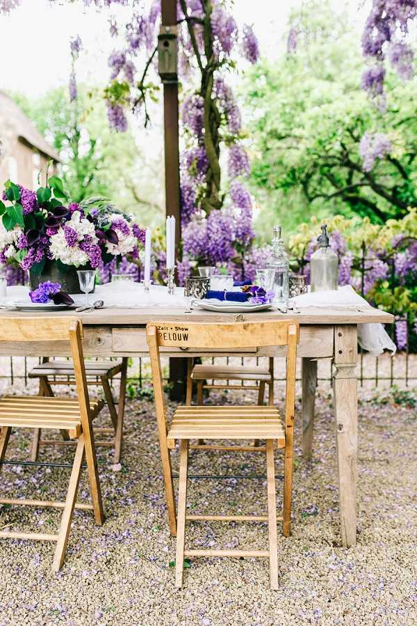 An outdoor wedding tablescape set on a gravel courtyard, featuring a rustic bare-wood farm table surrounded by lightweight natural wood folding chairs, one of which is branded with text reading 'Zelt + Show PELOUW.' The table is styled with a centerpiece of deep purple calla lilies, white and purple lilacs, and white hydrangeas in a dark metal vase, alongside tall white taper candles, silver candleholders, a glass soda siphon, and place settings with blue folded napkins on white plates. The decor palette combines deep purple, white, and silver in a relaxed rustic style. The shot is a medium wide angle taken at table level, capturing the full table setting in the foreground.