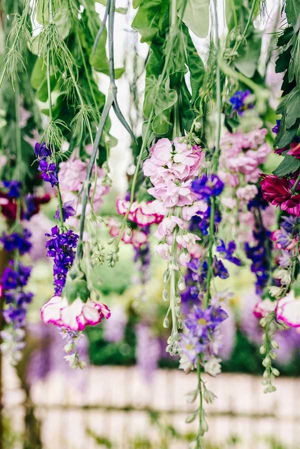 Close-up detail shot of a hanging floral installation used as wedding decor, photographed outdoors with a blurred garden background. The arrangement features cascading stems of purple and blue delphinium, light pink snapdragons, deep magenta ranunculus with white edges, and abundant green foliage including trailing leaves and tendrils. The color palette is a mix of purple, violet, soft pink, hot pink, and deep magenta against lush green stems. The installation appears to be suspended overhead, creating a ceiling or canopy effect, with a white fence structure visible and soft purple floral elements blurred in the background.