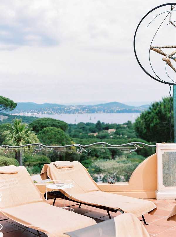 An outdoor terrace scene with two cream-colored sun loungers positioned on terracotta tile flooring, with a small white side table between them holding glassware. A decorative wrought-iron armillary sphere sculpture is visible in the upper right corner, and an ornate scrolled metal railing borders the terrace. The wide-angle shot looks out over an expansive view of forested hills and a bay with sailboats visible in the distance. No people or wedding party are present in this image. Potential venue feature image.