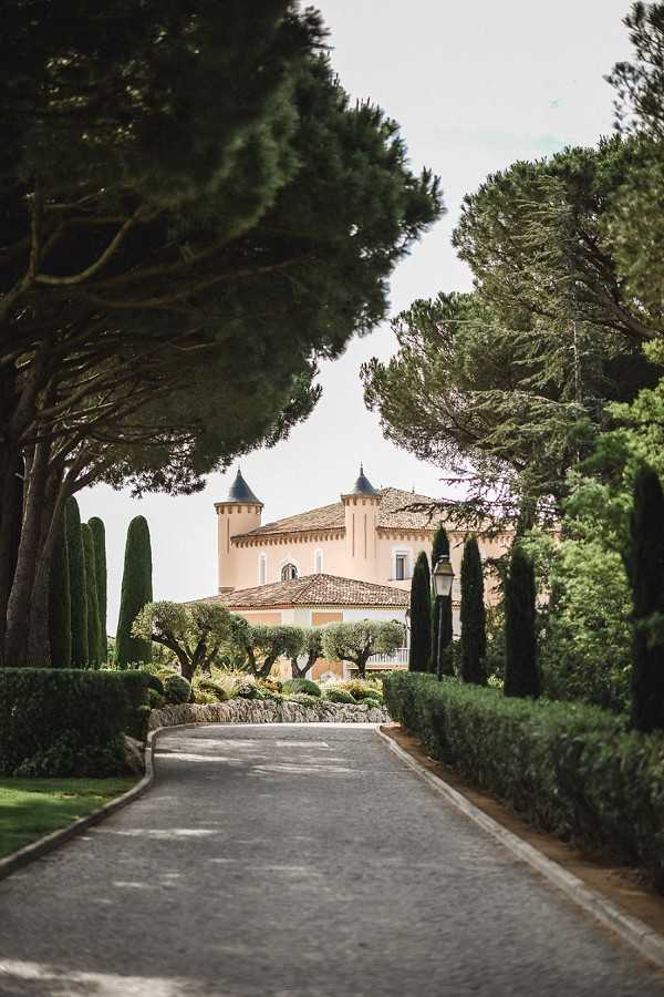 Wide shot of a paved driveway leading toward a peach-colored French chateau with two distinctive round towers topped with dark slate conical roofs and terracotta roof tiles. The approach is framed by tall columnar cypress trees, manicured hedgerows, and large umbrella pine trees on either side, with a classic black lantern post visible along the path. No people are present in the image. Potential venue feature image.
