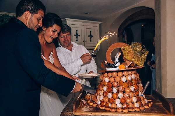 The bride and groom are cutting a traditional French croquembouche wedding cake, a tower of cream puffs, during an indoor reception. The croquembouche is topped with a classic bride-and-groom cake topper, decorative straw, and an active sparkler, while a man in a white shirt assists behind them. The bride wears a lace-detailed ivory gown and the groom a dark navy suit jacket. The setting appears to be inside a rustic stone-walled venue with an arched doorway and cream-painted cabinetry visible in the background, lit with warm ambient lighting. This is a medium close-up shot capturing the energy and movement of the cake-cutting moment.