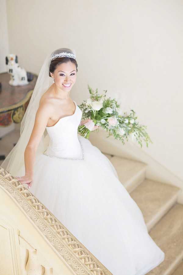 A bridal portrait taken indoors on an ornate staircase with cream-painted carved wood balustrade detailing, shot from a slightly elevated angle. The bride wears a strapless white ball gown with a beaded waist detail, a cathedral-length veil, and a crystal tiara, her dark hair pulled back neatly. She holds a loose, garden-style bouquet of white and blush roses with trailing greenery and ferns. The interior setting features light neutral walls and a small decorative side table with ceramic figurines visible in the background, suggesting a chateau or manor house interior.
