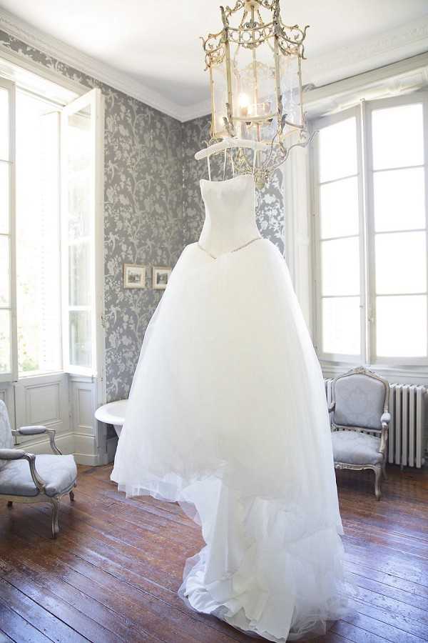 A getting-ready detail shot of a white strapless ball gown wedding dress with a structured sweetheart bodice and a voluminous tulle skirt with a ruffled hem, hung from an ornate gold lantern-style chandelier. The dress is displayed in a classic French interior room featuring grey damask wallpaper, dark hardwood floors, tall white-framed windows letting in natural light, a white radiator, and two Louis XVI-style upholstered chairs in pale grey. A delicate necklace is draped across the bodice of the dress. The wide shot captures the full dress against the period-appropriate décor, giving the image a classic, formal aesthetic consistent with a French chateau setting.
