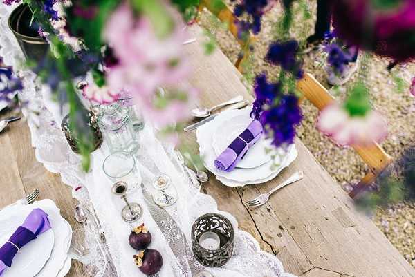 A close-up, slightly elevated detail shot of an outdoor wedding reception table set in a rustic boho style. The raw wooden farm table is dressed with a white lace table runner and set with white scallop-edged plates, silver cutlery, and purple folded napkins. Decorative elements include clear glass bud vases, an ornate dark metal votive candle holder, small silver candlestick holders, and fresh figs placed directly on the table. The floral arrangements visible in the foreground and background feature deep purple and violet blooms with greenery, softly blurred and framing the composition. The overall color palette centers on purple, white, and natural wood tones, consistent with a romantic boho outdoor styling.