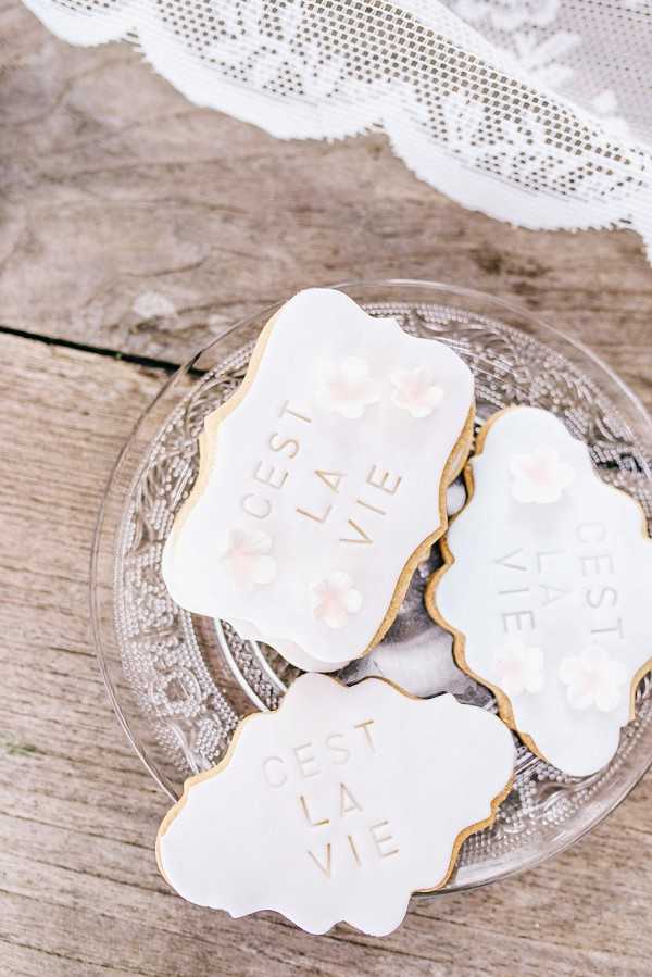 Close-up detail shot of three custom royal icing sugar cookies displayed on a clear pressed-glass plate, placed on a weathered wooden surface. The cookies are shaped in a decorative plaque/cartouche form with scalloped edges, covered in white fondant icing with gold lettering reading 'C'EST LA VIE' and small blush pink floral sugar decorations. A white lace ribbon or fabric trim is partially visible at the top of the frame, adding to the white and gold styling palette. The overall aesthetic is classic French-themed with a soft, white and blush decor palette.