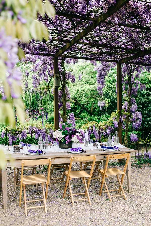 An outdoor reception table styled under a wooden pergola covered in cascading purple wisteria blooms, set on a gravel surface. The rustic wooden farm table is surrounded by natural wood folding chairs and set for approximately six guests, with gray plates, purple folded napkins, glassware, and taper candles in slim holders. The centerpiece features a low arrangement of white hydrangeas and purple florals in a dark vessel, with additional smaller floral accents and votives running along the table. The styling palette is purple and white with natural wood tones, giving an organic, garden-party aesthetic. Wide shot taken from slightly beyond the table, with wisteria-covered foreground framing the scene. Potential venue feature image.