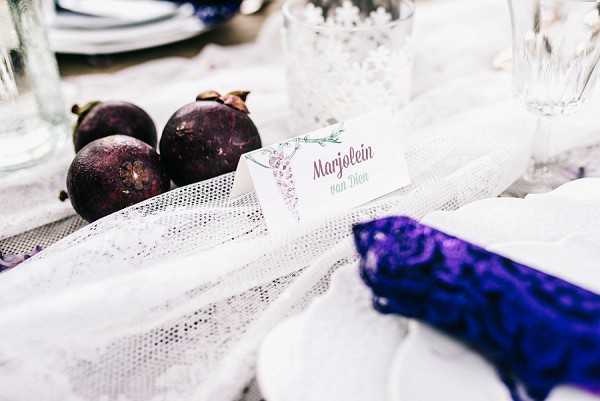 Close-up detail shot of a wedding reception tablescape featuring a place card reading 'Marjolein van Dien' with lavender-toned botanical printing. The table is styled with a white lace table runner, and deep purple-black figs are used as organic decorative elements alongside the place setting. A vivid royal blue lace napkin is folded and placed on a white plate to the right of the frame, anchoring the purple and white color palette. Glassware and additional place settings are softly visible in the background, suggesting a styled indoor reception.
