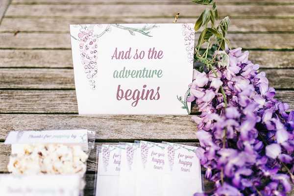A close-up detail shot of a wedding stationery and favor display arranged on a weathered wooden surface. A printed card reading 'And so the adventure begins' features watercolor wisteria illustrations in purple and green. Beside it, small labeled favor packets reading 'for your happy tears' are fanned out, alongside a clear bag of popcorn tagged with 'thanks for popping by.' Fresh purple wisteria blooms are placed to the right of the display. The overall decor palette centers on purple, mauve, and green, consistent with a garden or boho-inspired wedding styling theme.
