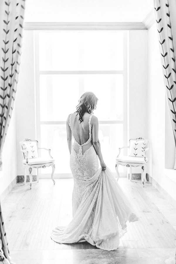 This is a black-and-white portrait of a bride standing indoors in front of a large window, shot from behind. The image features bright, high-key lighting from the window, creating a light, airy contrast against the wooden floor and white walls. The bride is wearing a form-fitting lace gown with a deep open back, thin straps with lace detailing along the neckline, and a flowing train that she gently holds to one side. Her dark hair is loosely styled and worn down. Two ornate French-style white armchairs with patterned upholstery are positioned symmetrically on either side of the window, and printed curtains with a leaf motif frame the scene, suggesting a classic, refined interior setting.