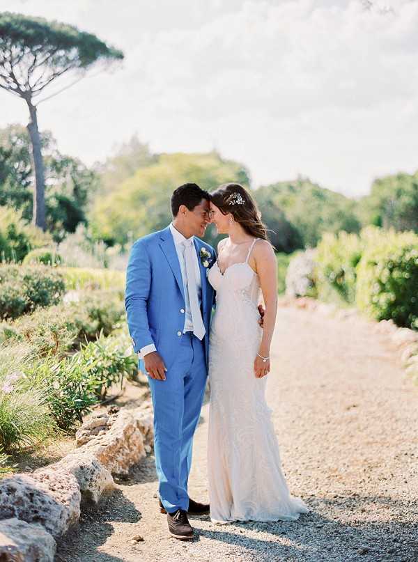 A couple portrait taken outdoors on a gravel path surrounded by Mediterranean garden landscaping, with tall umbrella pine trees visible in the background. The groom wears a cornflower blue suit with a light blue tie and a small blue floral boutonniere, while the bride wears a fitted ivory lace gown with spaghetti straps and a sweetheart neckline, accessorized with a delicate hair piece and bracelet. The two stand close together with foreheads nearly touching, smiling at each other in a candid, relaxed pose. The image is a medium-length portrait shot with warm natural afternoon light and a soft, film-style finish.