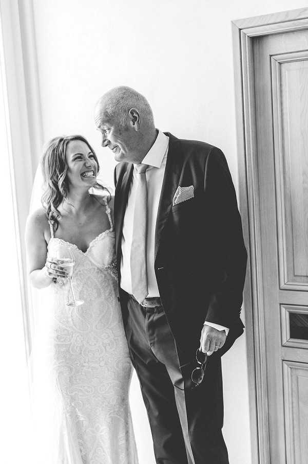A black-and-white portrait of a bride and an older man, likely her father, sharing a candid laugh together indoors against a bright white wall near a wooden door. The bride is wearing a fitted lace wedding dress with spaghetti straps and a sweetheart neckline, and is holding a champagne flute in one hand. The man is dressed in a dark suit with a light-colored tie and a patterned pocket square, and is holding a pair of sunglasses in his hand. The image is shot in a mid-length portrait style with high contrast and bright, airy tones.