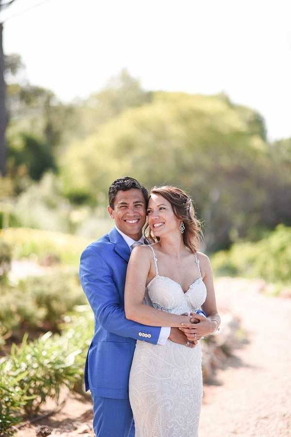 A couples portrait taken outdoors on a gravel path surrounded by garden greenery in bright natural daylight. The groom stands behind the bride with his arms wrapped around her waist, both smiling broadly at the camera. He wears a cobalt blue suit with a white dress shirt, and she wears a fitted ivory lace gown with spaghetti straps, a sweetheart neckline, and chandelier earrings with her hair loosely styled. The shot is a medium portrait framed from roughly the knees up, with the background softly blurred.