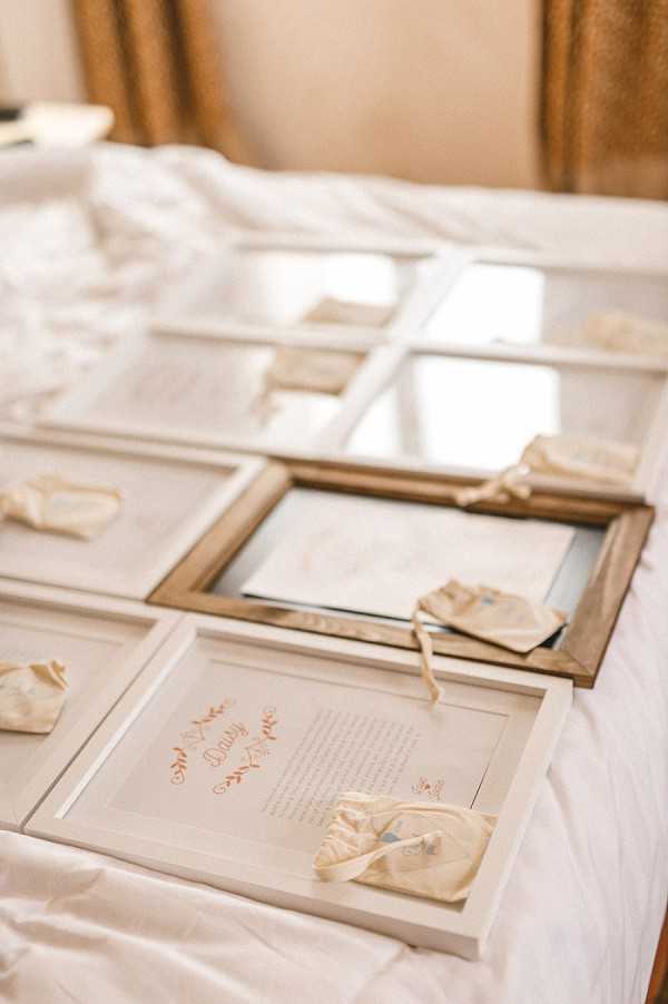 Close-up detail shot of wedding stationery or keepsake frames laid out on a white satin-draped surface, likely during a getting-ready or preparation phase. Multiple white and gold-framed pieces are displayed, each appearing to contain handwritten or printed text with coral/rose-colored calligraphy headings — the closest frame is legible with the name 'Daisy' and decorative botanical motifs. Each frame has a small ivory ribbon or fabric bow attached as a decorative accent. The overall palette is ivory, white, and gold with coral script details, suggesting a classic and refined styling approach. The background shows gold drapery, indicating an indoor setting.