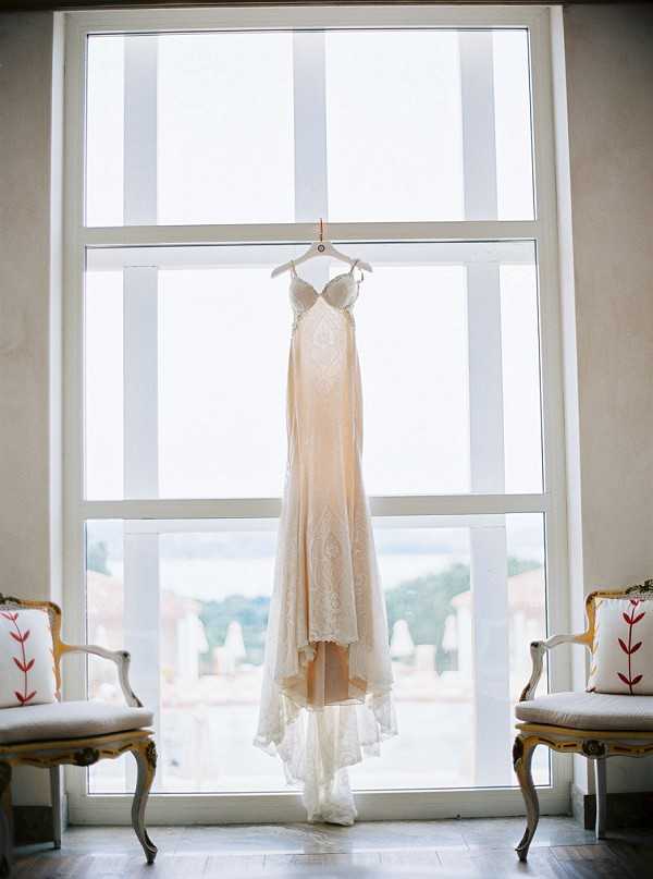A getting-ready detail shot of a bridal gown hanging from a hanger in front of a large multi-pane window, backlit by natural light. The dress is a champagne and ivory spaghetti-strap slip-style gown with lace overlay detailing on the bodice and hem, featuring a slight train. On either side of the window sit two gold-framed French-style armchairs with light grey upholstery and white cushions printed with a red botanical motif. The wide shot composition frames the dress symmetrically between the two chairs, with a rooftop view and distant treeline visible through the window.