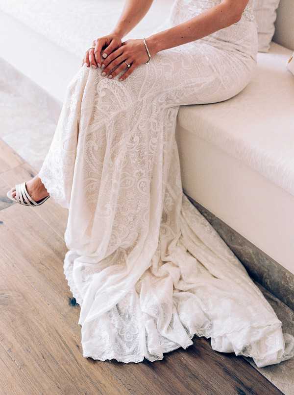 A close-up detail shot of a bride seated on a light-colored upholstered bench or sofa in an indoor setting with wood flooring. The image focuses on her ivory lace wedding dress, which features an intricate all-over paisley and scroll lace pattern with a flowing train pooling on the floor. Her hands rest in her lap, showing a delicate pearl or diamond bracelet and an engagement ring, with neatly manicured nails. She wears white strappy heeled sandals visible at the lower left of the frame.