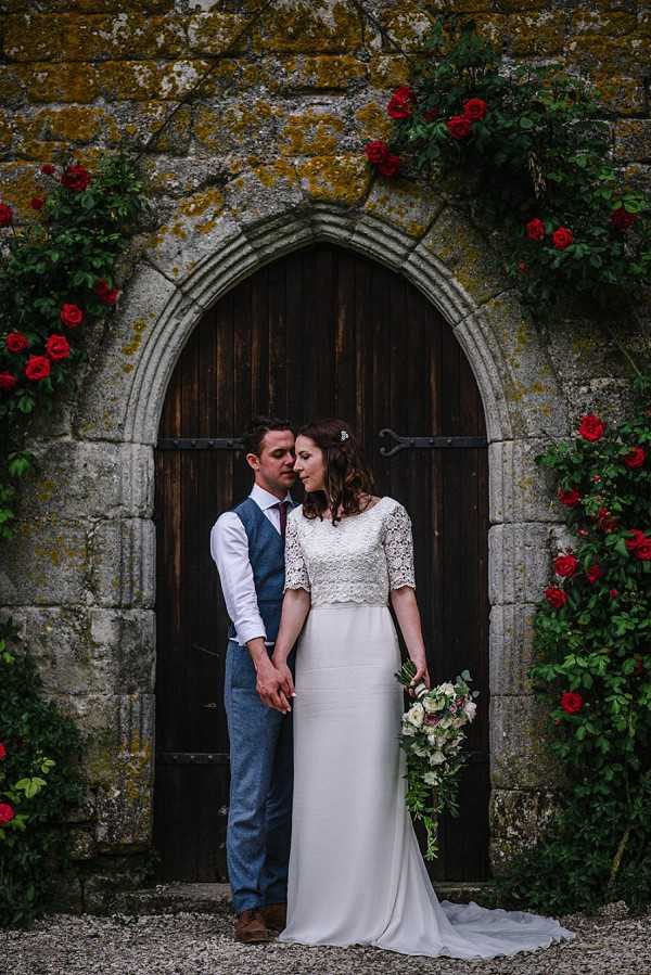 A couple portrait taken outdoors in front of a large arched gothic wooden door set into a stone wall covered with climbing red roses. The bride wears a two-piece ivory gown with a short-sleeved lace crop top and a plain fitted skirt with a small train, and holds a loose, trailing bouquet of cream and blush blooms with greenery. The groom wears a blue tweed waistcoat, white shirt, burgundy tie, and blue trousers with brown shoes. The two stand close together holding hands, with the groom leaning his head toward the bride's. The composition is a full-length portrait shot with the doorway and climbing roses framing the couple symmetrically.