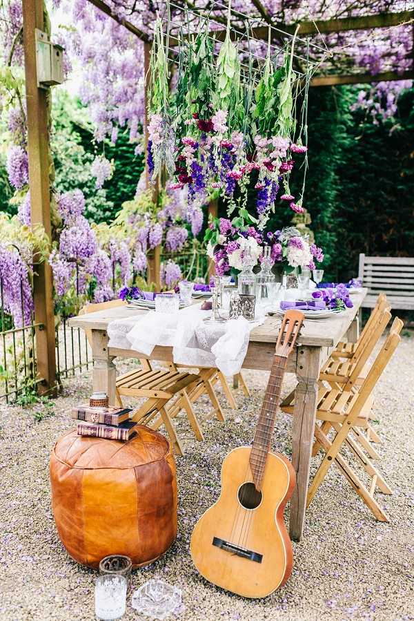 An outdoor boho-style wedding reception tablescape styled under a wooden pergola covered in blooming wisteria. The rustic wooden dining table is set with a white lace table runner, clear glassware, purple folded napkins, and a centerpiece of purple, lavender, and white blooms including what appear to be delphiniums and hydrangeas. A hanging floral installation suspended from the pergola features cascading greenery mixed with deep burgundy, purple, and pink blooms. In the foreground, a tan leather moroccan pouf sits beside a stack of vintage books, a small candle, and an acoustic guitar leaning against the table. Wooden folding chairs surround the table, and the gravel ground has scattered wisteria petals. Wide editorial shot showcasing the full table setting and decor.