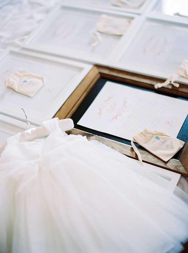 A detail flat-lay shot showing wedding or event stationery and accessories arranged on a surface. Multiple white-framed cards or menus with pink handwritten-style calligraphy text are displayed alongside small cream-colored fabric favor pouches tied with ivory ribbon. In the foreground, a miniature white tulle flower girl dress hangs from a small hanger, featuring a fitted bodice with a full layered tulle skirt. A warm wood-toned frame holds one of the printed cards at center. The overall palette is ivory, cream, and soft blush, suggesting a classic or romantic styling theme.
