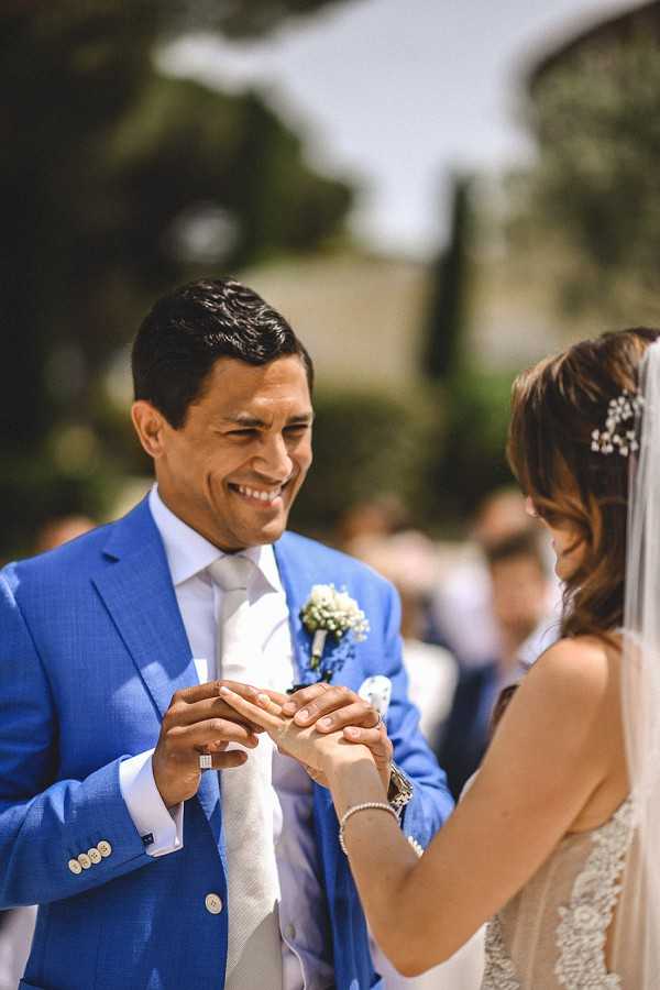 The ring exchange moment during an outdoor ceremony, with the groom placing a ring on the bride's finger. The groom wears a cobalt blue suit with a light grey tie and a white baby's breath boutonniere, and is smiling broadly. The bride, partially visible from behind, wears a lace sleeveless gown, a sheer veil with a small floral hair accessory, and a delicate pearl bracelet. The background shows a blurred group of seated guests and what appears to be a stone building partially visible through trees, suggesting a chateau or villa setting. This is a close-up portrait shot with shallow depth of field, keeping the focus on the couple's hands and the groom's expression.