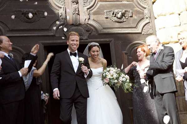 A couple exits through the large ornate dark wooden doors of a stone chapel or church as guests toss white flower petals in celebration. The groom wears a classic black tuxedo with a white boutonniere and black bow tie, while the bride wears a strapless white ballgown with a cathedral veil and a tiara, carrying a bouquet of blush and ivory roses with greenery. Approximately six guests are visible on either side, dressed in formal attire, actively throwing petals. The shot is a medium wide portrait taken at the church entrance, capturing the joyful recessional moment.