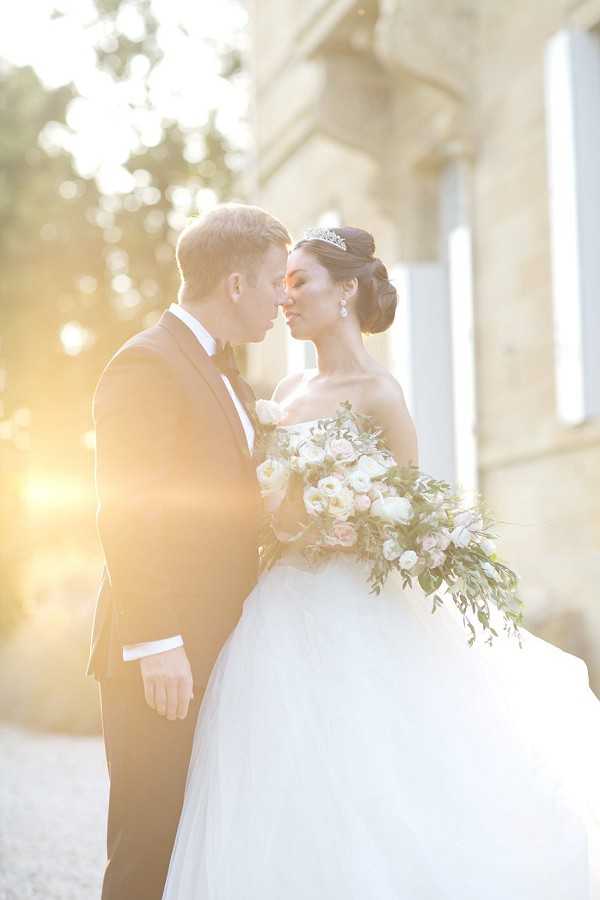 A couple portrait taken outdoors in front of a classical stone building, shot with strong golden-hour backlight creating a warm lens flare on the left side of the frame. The groom wears a black tuxedo with a bow tie, and the bride wears a strapless white ballgown with a full tulle skirt; her dark hair is styled in an updo with a diamond tiara and drop earrings. She holds a large cascading bouquet of ivory ranunculus, blush roses, and trailing greenery. The two are facing each other with foreheads nearly touching in a close romantic pose. The composition is a mid-length portrait with a shallow depth of field that softly blurs the architectural background.
