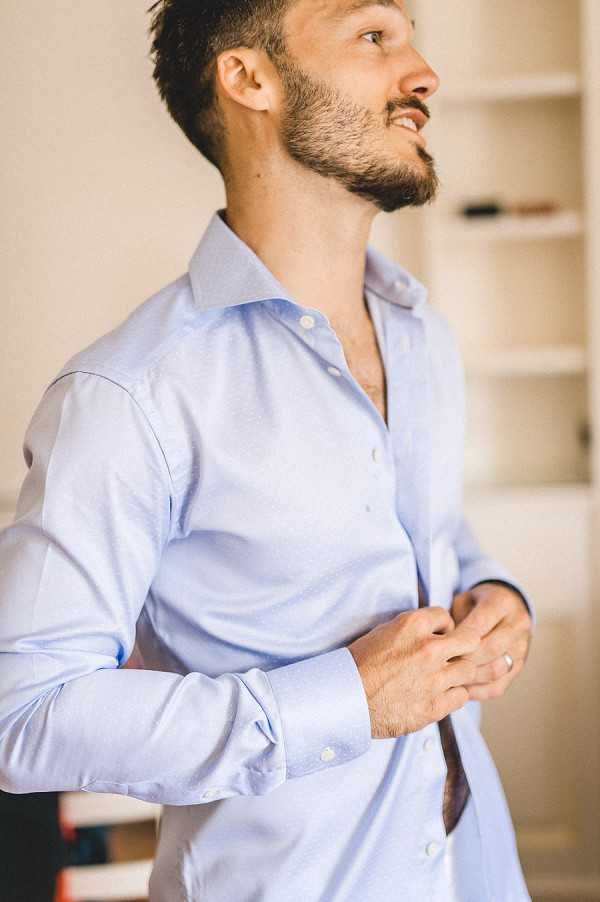 A groom getting ready indoors, buttoning up a pale blue dress shirt while looking upward with a slight smile. He wears a wedding band and has short dark hair and a trimmed beard. The background shows a bright, simply furnished room with white built-in shelving. The shot is a close-up portrait cropped at the torso, with soft natural light.