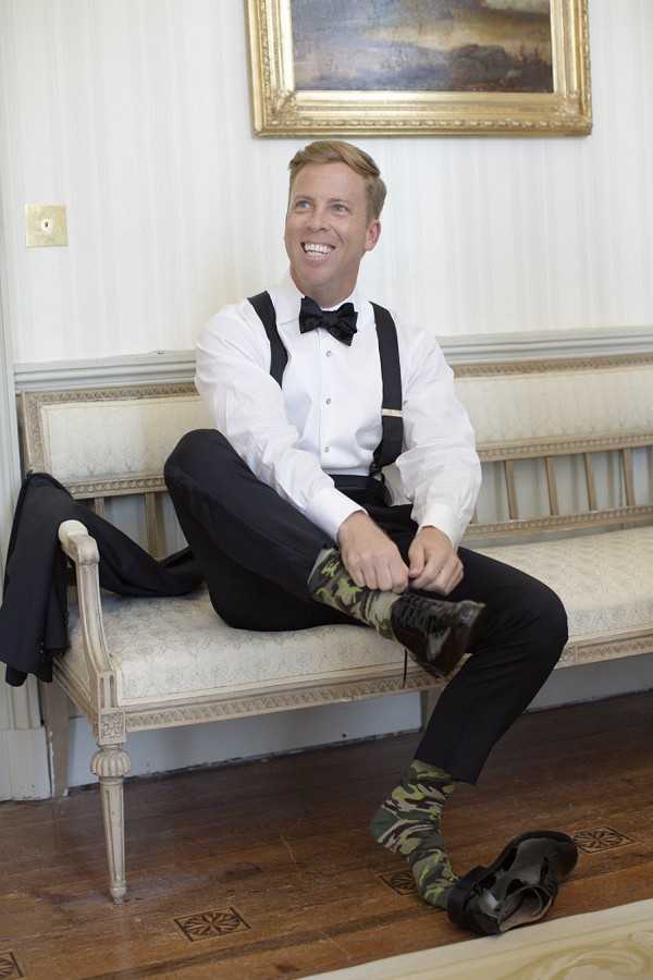 A groom is photographed getting ready indoors, seated on an upholstered cream-colored bench with carved wooden detailing, laughing and looking to the side while putting on his shoes. He is wearing a white dress shirt, black bow tie, black suspenders, and black trousers, with a notable intentional style choice of green camouflage-patterned socks. His black dress shoes are on the floor in front of him, and his black jacket is draped over the arm of the bench. A gold-framed oil painting hangs on the striped wallpapered wall behind him. The shot is a full-length portrait taken in what appears to be a formal interior room of a chateau or manor house.