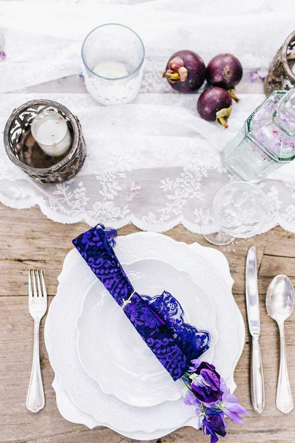 A close-up flat lay of a wedding reception place setting styled on a rustic bare wood table. The place setting features stacked white scalloped-edge plates with a deep royal blue lace-wrapped napkin roll tied with gold twine and accented with a small purple and lavender sweet pea bloom. A white floral lace table runner runs down the center of the table, flanked by silver cutlery including a fork, knife, and spoon. Decorative elements include a silver filigree votive candle holder with a white tea light, a light blue textured drinking glass, a clear faceted glass vessel, and a cluster of deep purple mangosteens used as organic decor. The overall color palette pairs white and silver with rich purple and royal blue tones in a boho-romantic styling.