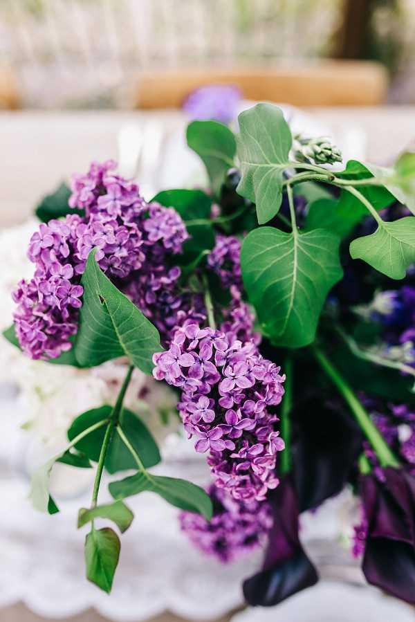 Close-up detail shot of a floral arrangement featuring purple lilac clusters and deep purple calla lilies with large green leaves, set against what appears to be a white lace or broderie anglaise table linen. The composition is tightly framed with a shallow depth of field, keeping the foreground lilacs in sharp focus while the background, which includes hints of additional purple florals and a wooden chair, blurs softly. The color palette is rich purple and green, suggesting a bold, garden-inspired floral design scheme.