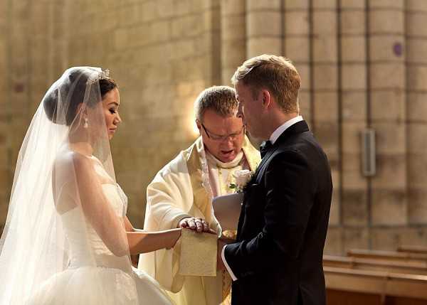 A wedding ceremony ring exchange is taking place inside a stone church or cathedral, with warm light filtering in from the side. The bride wears a white ball gown with a long cathedral veil and a small tiara, while the groom is dressed in a black tuxedo with a black bow tie. A priest in cream and gold liturgical vestments reads from a book while holding the couple's joined hands. The composition is a medium portrait shot focused on the three central figures, with wooden church pews visible in the soft-focus background.