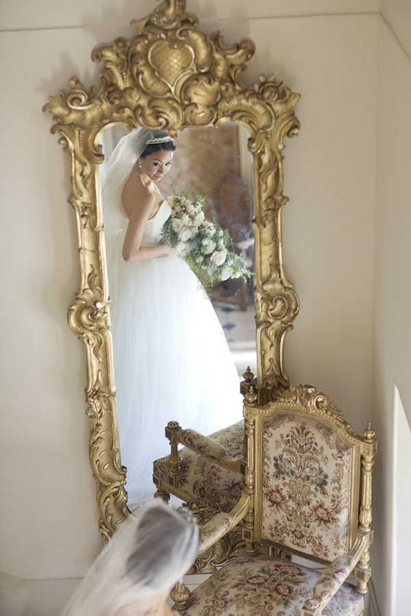 A bridal portrait captured as a reflection in a large ornate gold-framed mirror with elaborate Rococo-style carved detailing. The bride wears a full-skirt white ballgown with a long veil and a tiara, and holds a loose bouquet of blush pink roses and lush greenery. The image is composed from a slight angle, with the bride's veil visible in the foreground at the bottom of the frame, adding depth. A gilded antique chair upholstered in floral tapestry fabric sits beside the mirror, reinforcing the classic French interior styling. The setting appears to be an indoor getting-ready room with pale walls, consistent with a chateau-style venue.