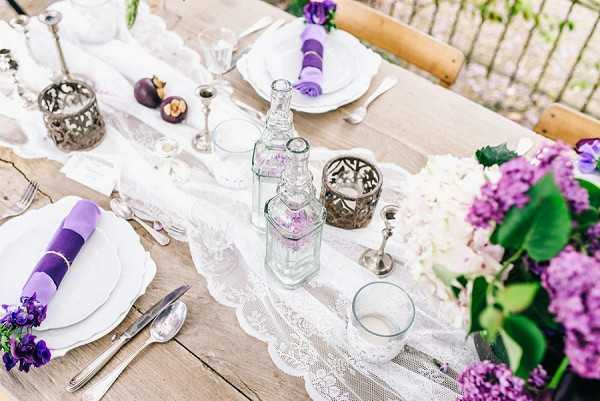 A close-up detail shot of an outdoor wedding reception tablescape set on a rustic wooden farm table. The table is dressed with a white lace runner and features a purple and white color palette, with bright purple linen napkins folded and placed on white ceramic plates, silver cutlery, and crystal glassware. Decorative elements include ornate silver lanterns and candle holders, clear glass bottles used as bud vases, and clusters of purple and white lilac blooms with dark purple sweet peas arranged along the table edge. A few dark-hued decorative fruits are also visible as accents. The overall styling is romantic and vintage-inspired, combining lace, silver metalwork, and fresh florals.