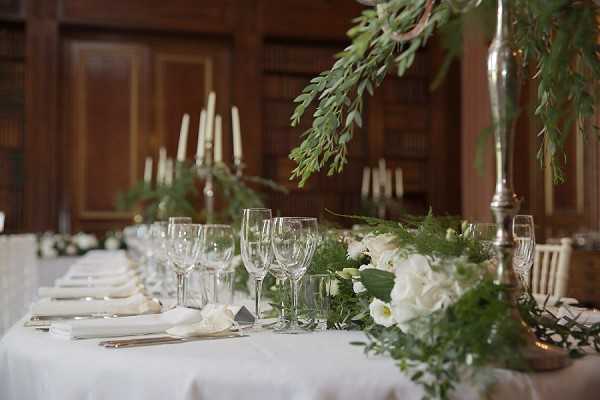 Close-up detail shot of a wedding reception tablescape set in a wood-paneled indoor room with built-in bookshelves visible in the background, suggesting a library or study setting. The long table is dressed in a white linen cloth and styled with a lush greenery runner featuring white roses and ranunculus, crystal wine glasses, white folded napkins, and silver candelabras holding ivory taper candles. A tall silver pedestal vase on the right side holds trailing greenery that cascades over the table. The overall decor palette is white, ivory, and green with silver metallic accents, creating a classic and refined aesthetic.