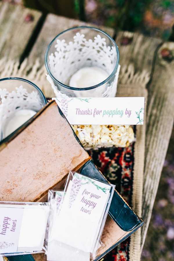 Close-up detail shot of wedding favor and guest amenity station arranged on a rustic wooden surface with a small patterned textile underneath. The display includes two glass tumblers wrapped with white laser-cut lace paper collars containing candles, a bag of popcorn labeled 'Thanks for popping by' in purple script with a green floral motif, and several small clear plastic packets of tissues labeled 'for your happy tears' in matching purple and green botanical typography. Vintage hardcover books in brown and dark blue are used as props to elevate the items. The styling is boho-rustic with a botanical theme, using a consistent purple and green color palette across the printed signage and packaging.
