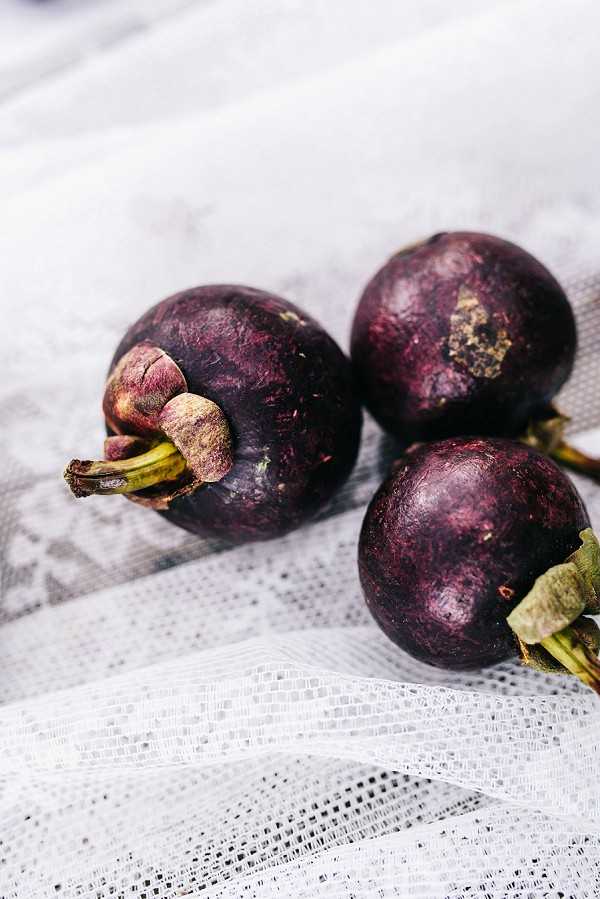 This image does not appear to be a wedding photograph. It shows three mangosteens — deep purple tropical fruits with green stems — arranged on a white mesh or tulle fabric. The shot is a close-up detail image with shallow depth of field. This does not contain any wedding-related content such as a couple, ceremony, reception, decor, or attire.