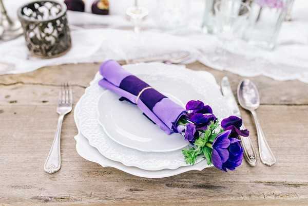 Close-up detail shot of a wedding table place setting on a rustic bare wood table. The setting features stacked white embossed ceramic plates with silver flatware on either side, and a rolled napkin in layered lavender and deep purple secured with a gold band and adorned with deep violet anemones and green foliage. In the soft-focus background, a dark ornate metal votive candle holder, a wine glass, and a pale linen table runner are partially visible. The decor palette centers on purple tones ranging from soft lavender to deep violet, suggesting a romantic, slightly bohemian styling direction.